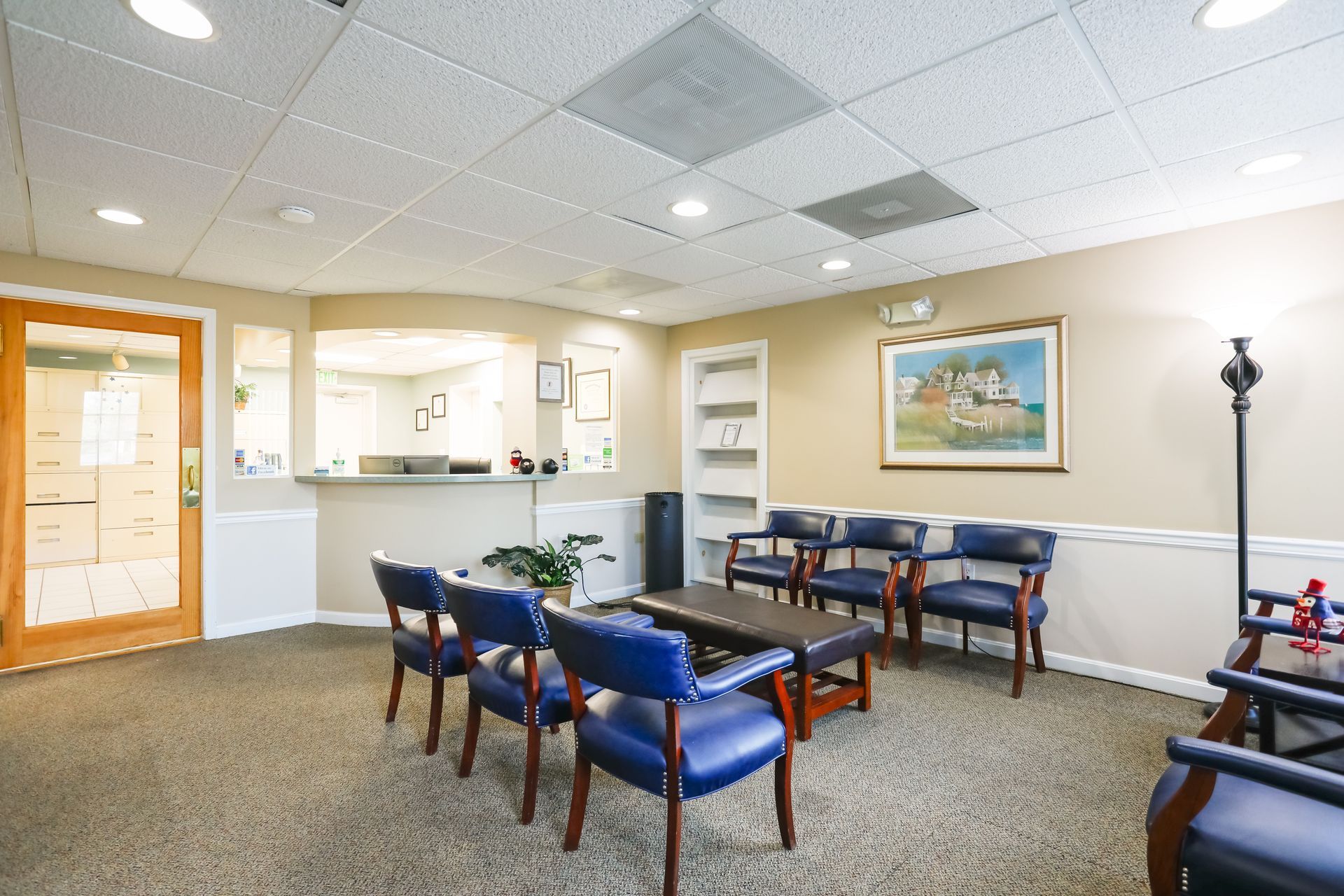 Empty waiting room with blue chairs, a small table, reception window, and framed wall art.