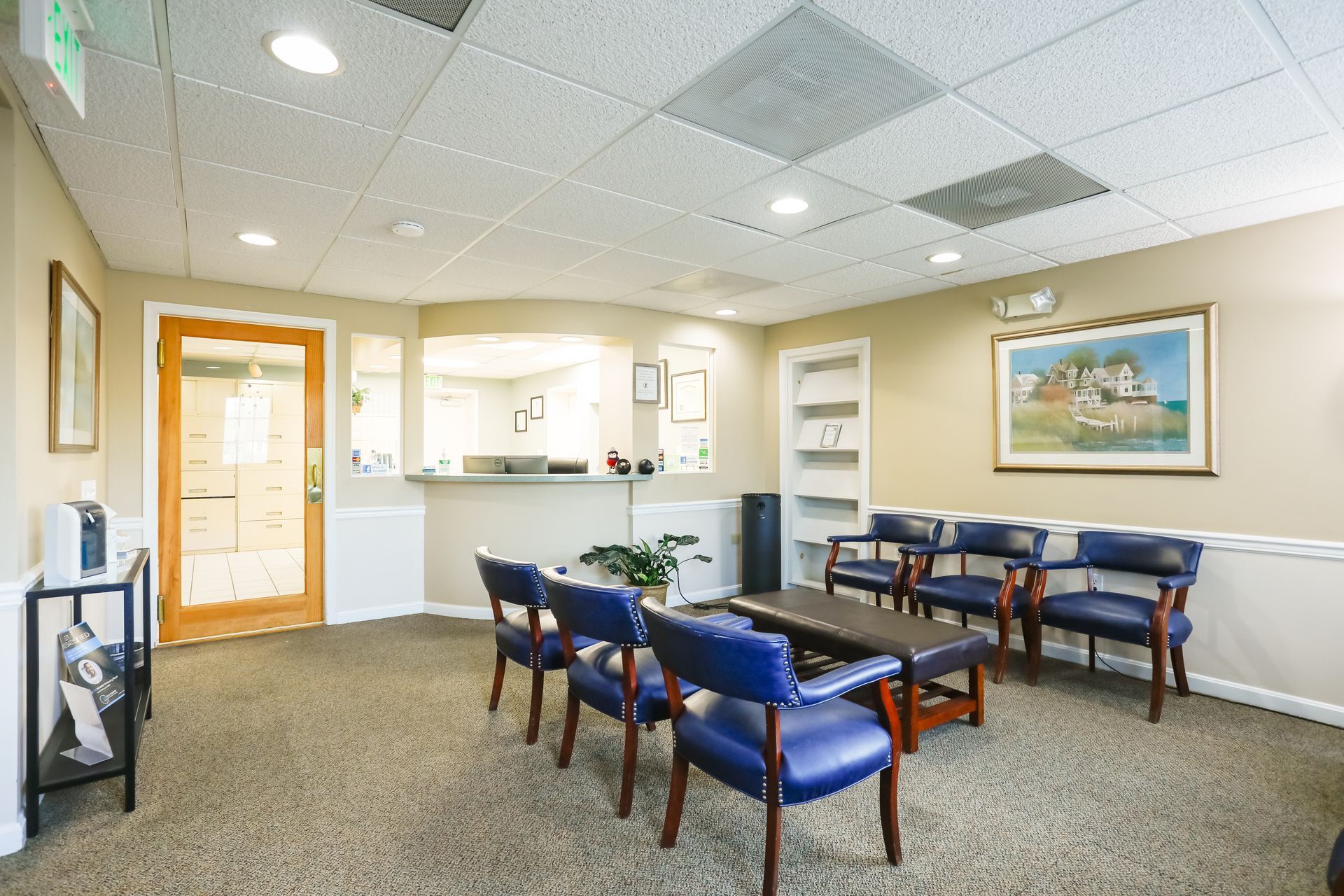 Empty waiting room with blue chairs, coffee table, wall art, and a glass entrance door.