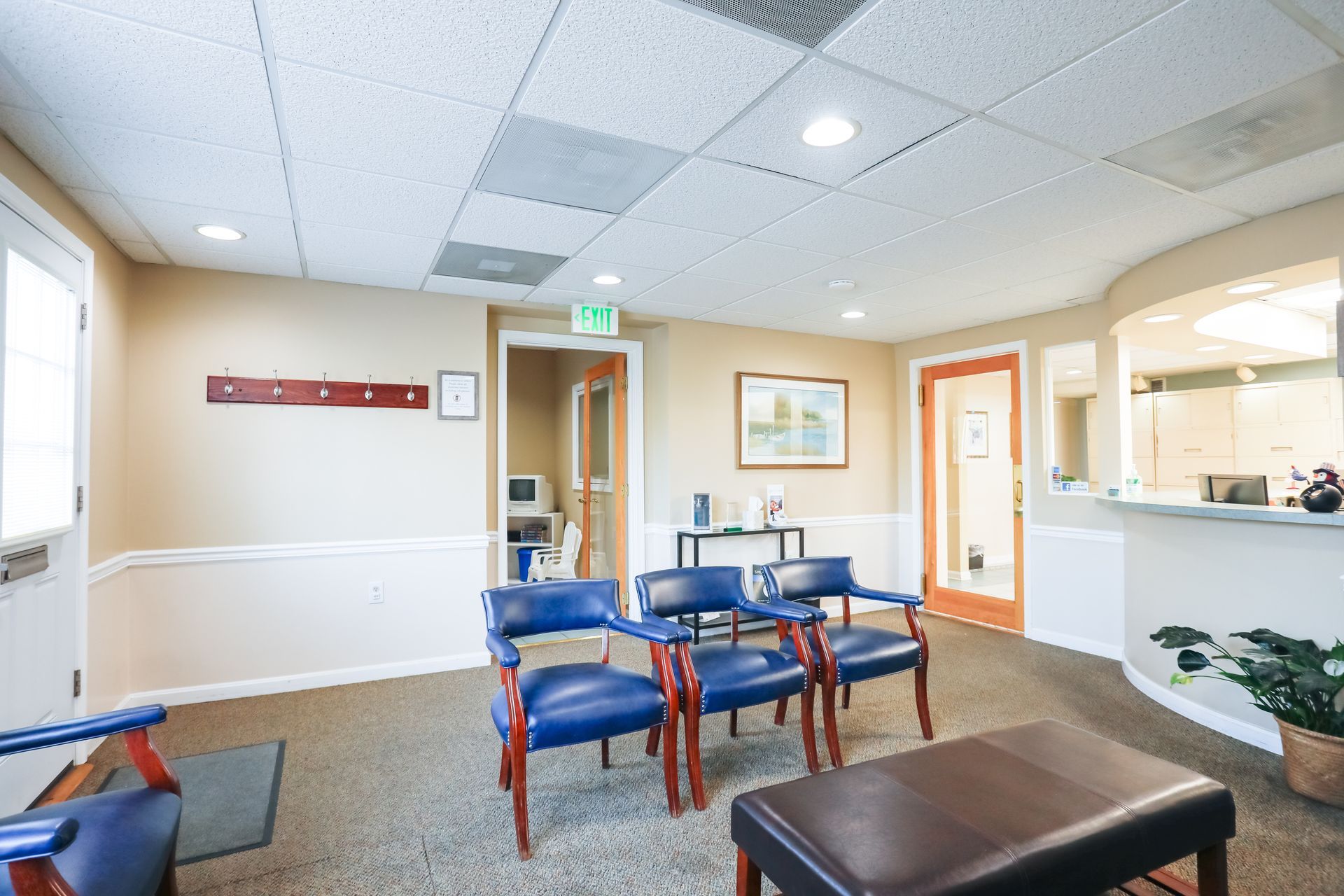 Empty medical waiting room with blue chairs, brown ottoman, reception desk, and hallway doors