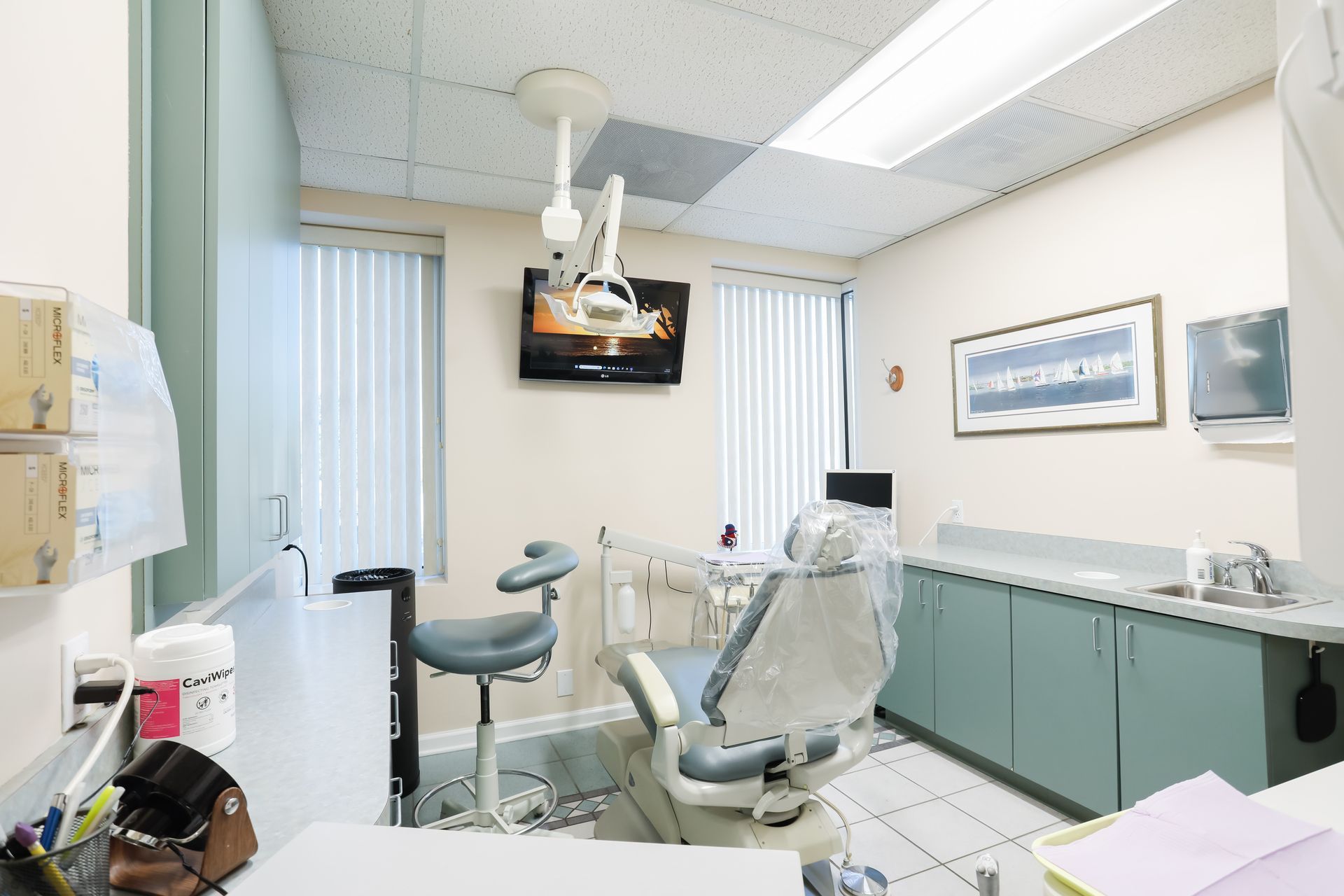 Dental exam room with chair, overhead light, cabinets, sink, and wall-mounted monitor.