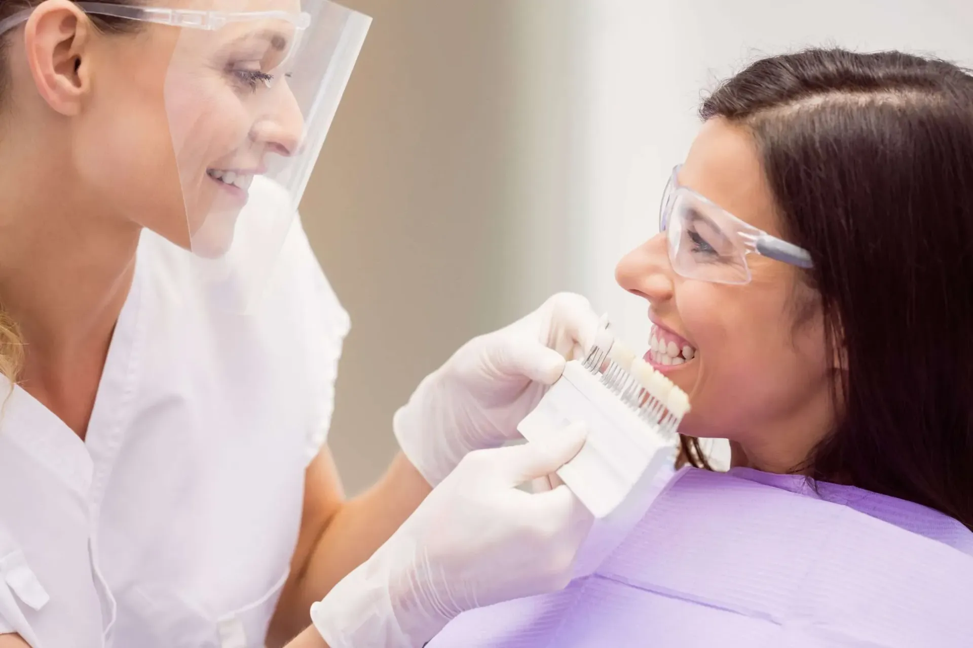 Dentist checking a young patient’s teeth while she smiles in a purple chair, both wearing face shields