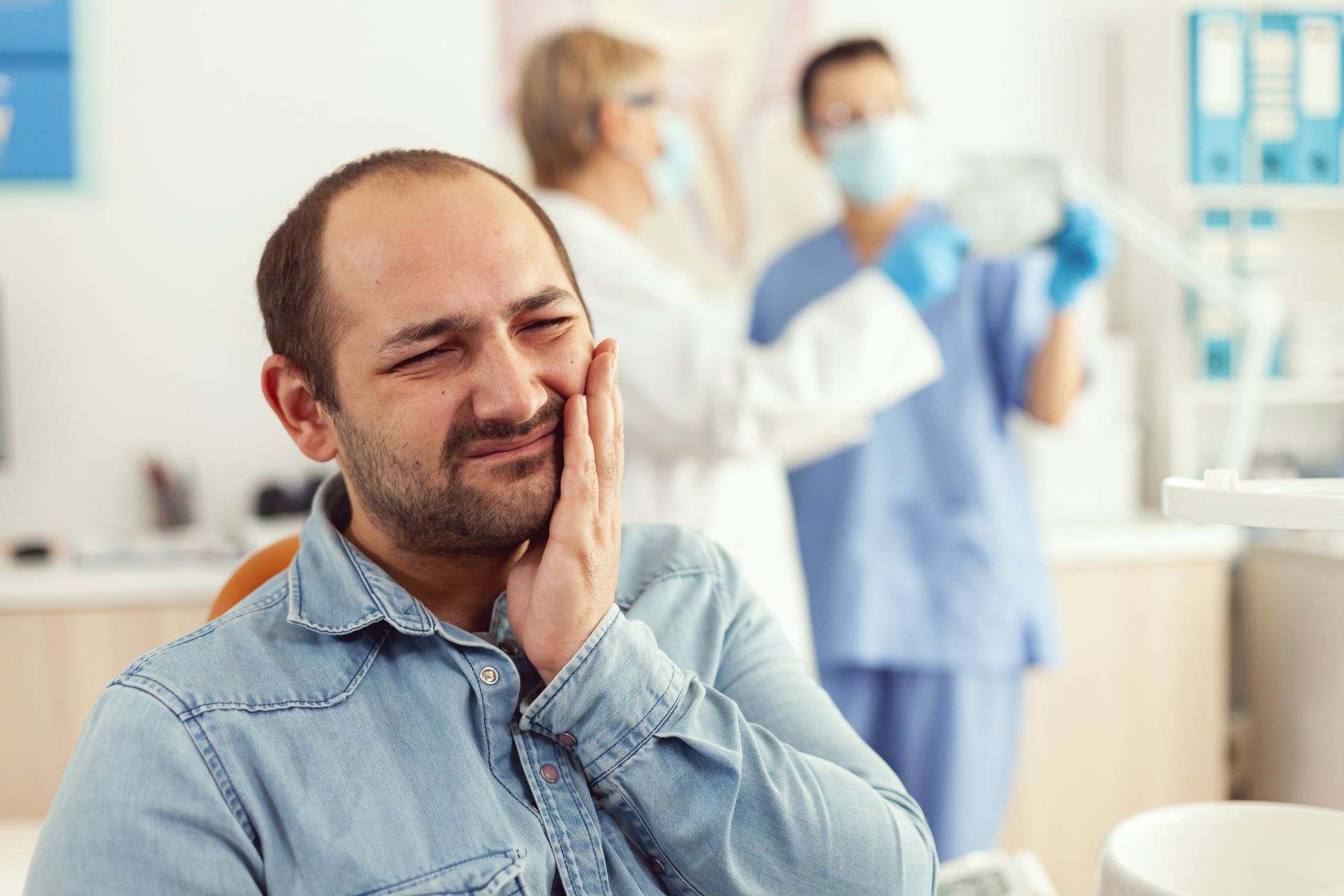Man holding his cheek in at a dental practice, grimacing in pain while staff stand in the background