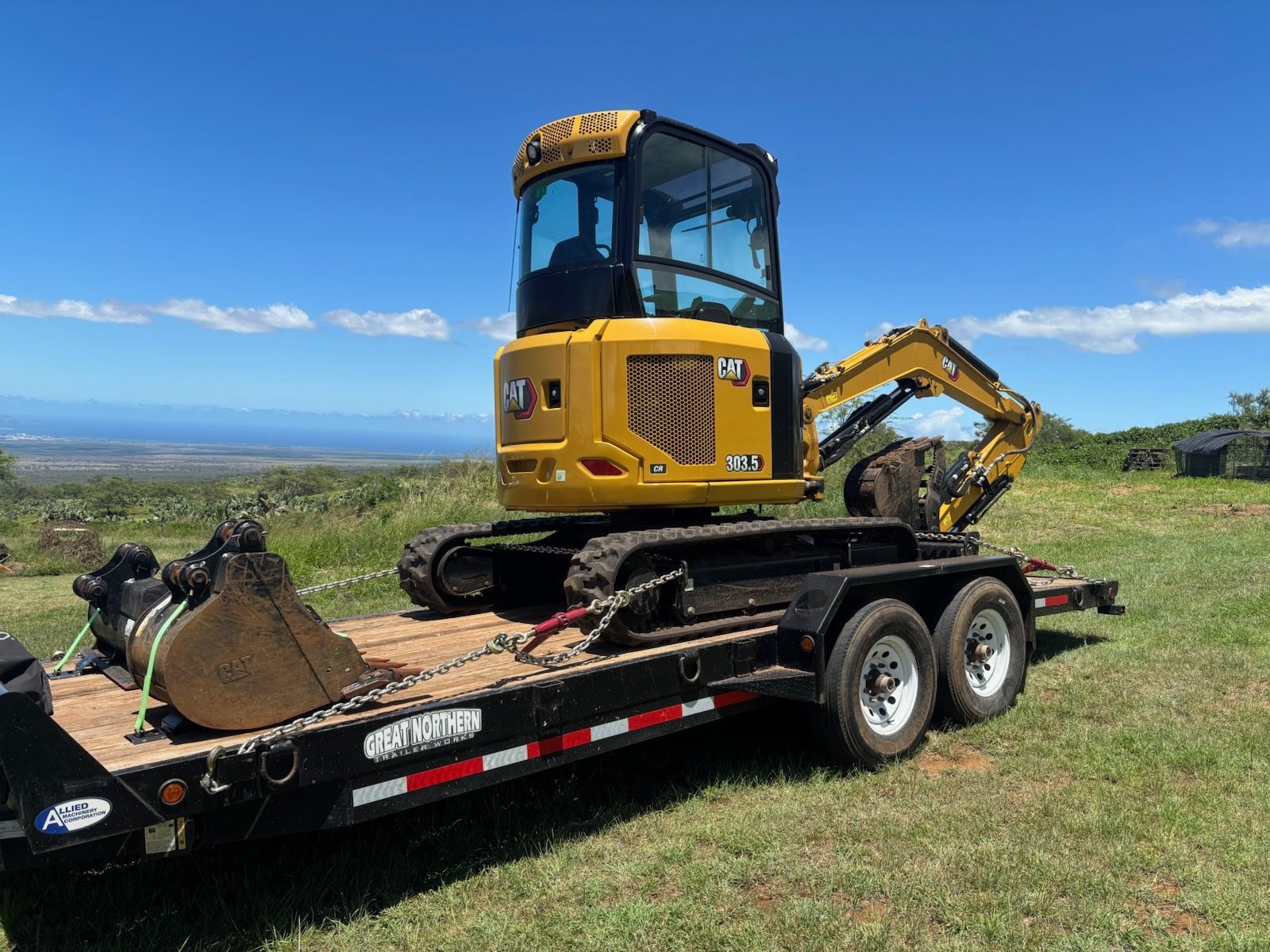 Yellow mini excavator on a black trailer in a grassy field under a blue sky.