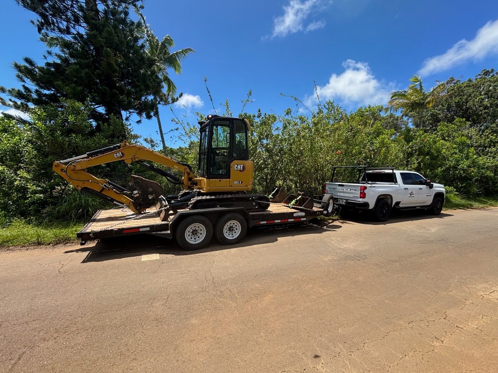 Yellow excavator on a flatbed trailer hitched to a white pickup on a dirt road under a blue sky.
