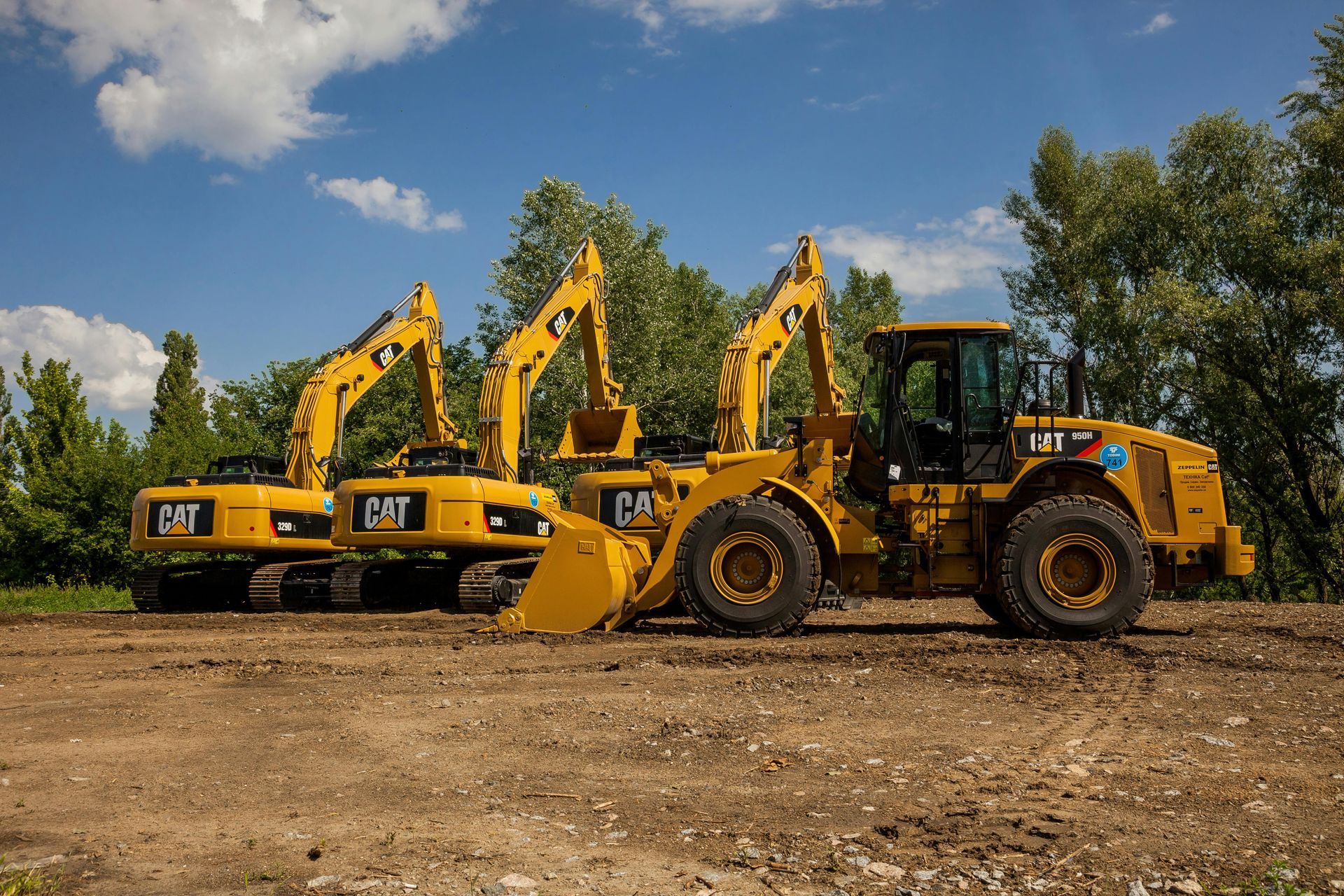 Three yellow Caterpillar construction vehicles parked on a dirt lot against a backdrop of trees and a blue sky.