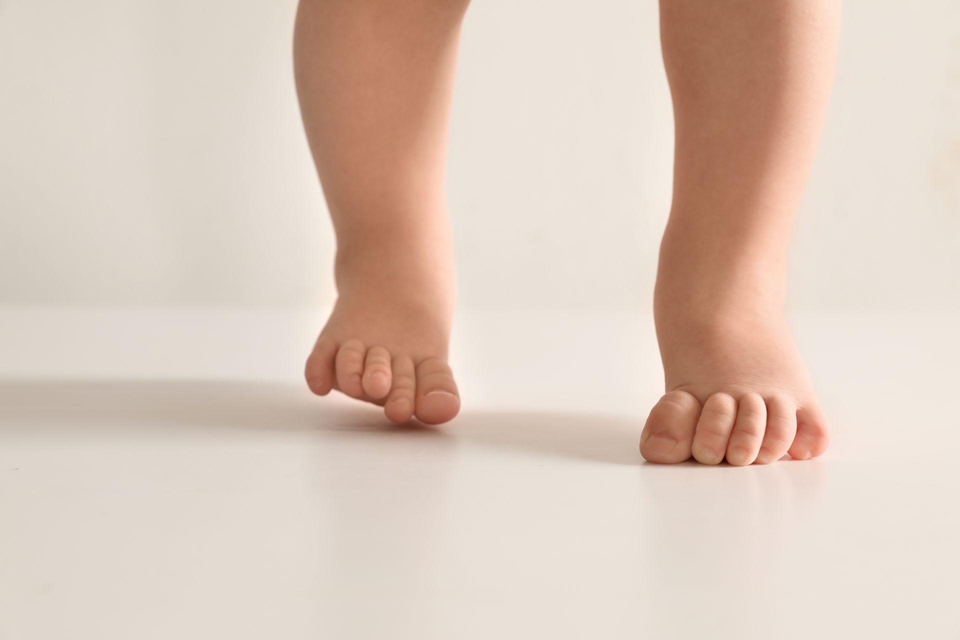 Baby's bare feet taking first steps, toes curled, on a white surface.