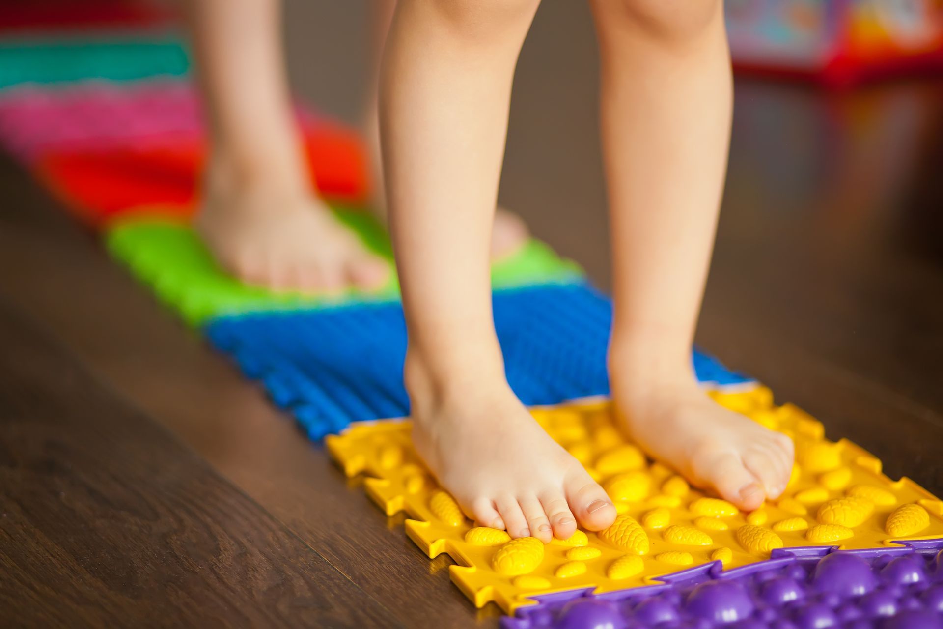 Child's bare feet walking on a colorful textured massage mat on a wooden floor.