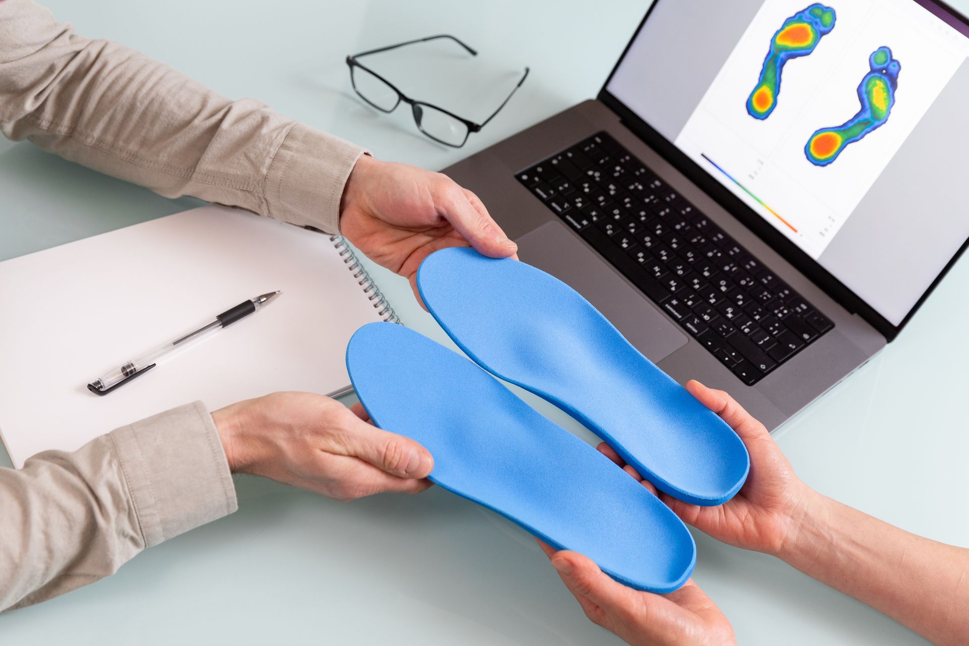 Hands holding blue orthotic insoles over a laptop displaying foot pressure analysis.