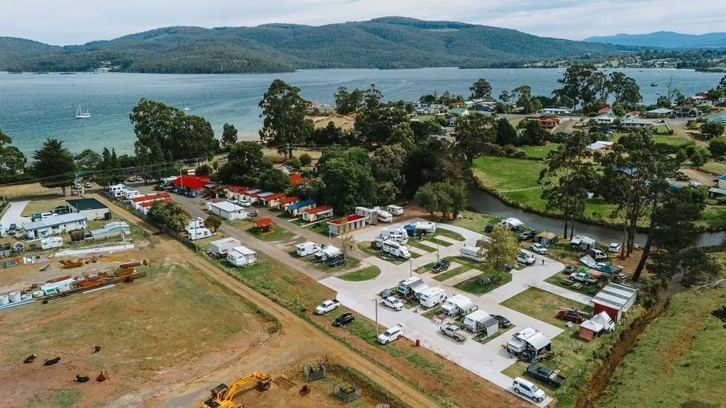 Aerial view of a lakeside campground with tents, vehicles, and trees beside water and hills. — Dover Caravan Park in Dover, TAS