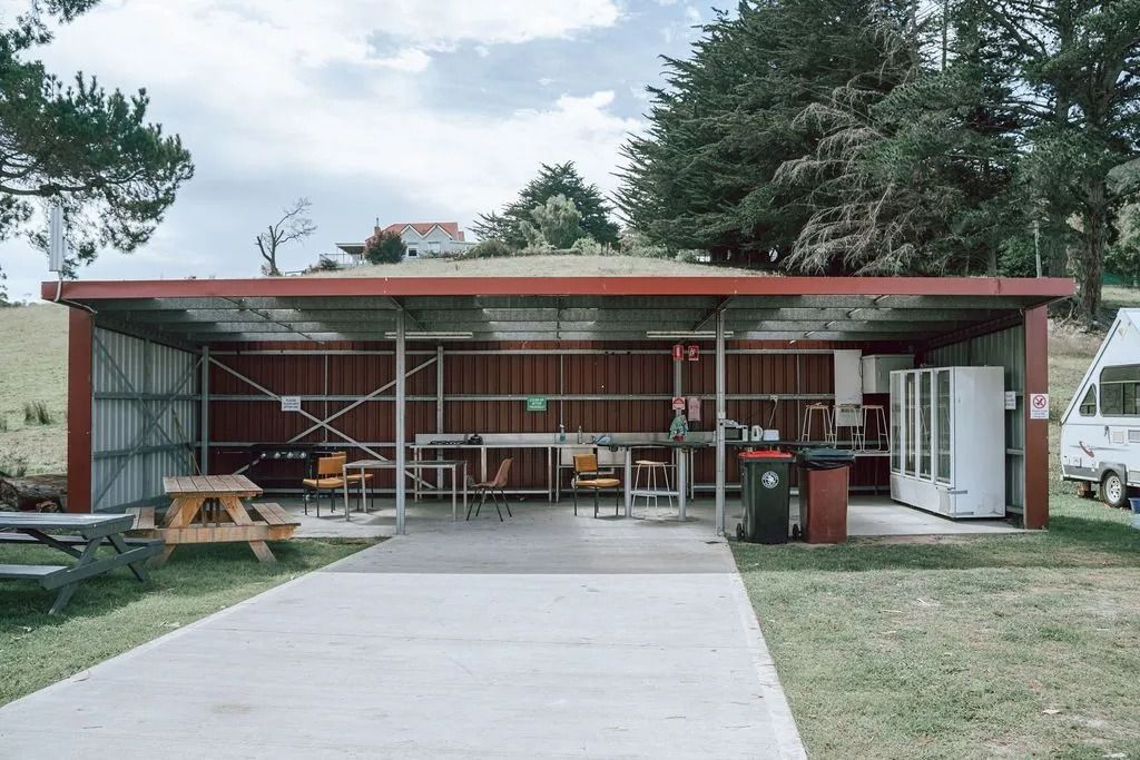 Open-air red-roof pavilion with tables, benches, and a central walkway in a grassy yard — Dover Caravan Park in Dover, TAS