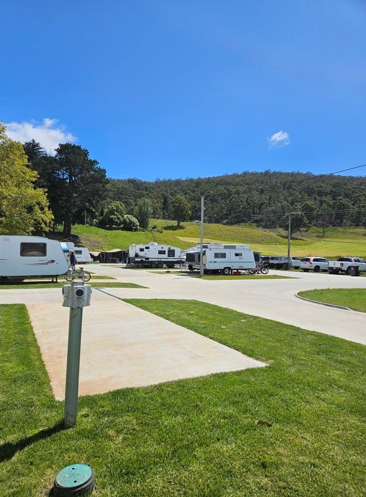 RV campers parked on a grassy campground under a blue sky, with hills in the background — Dover Caravan Park in Dover, TAS