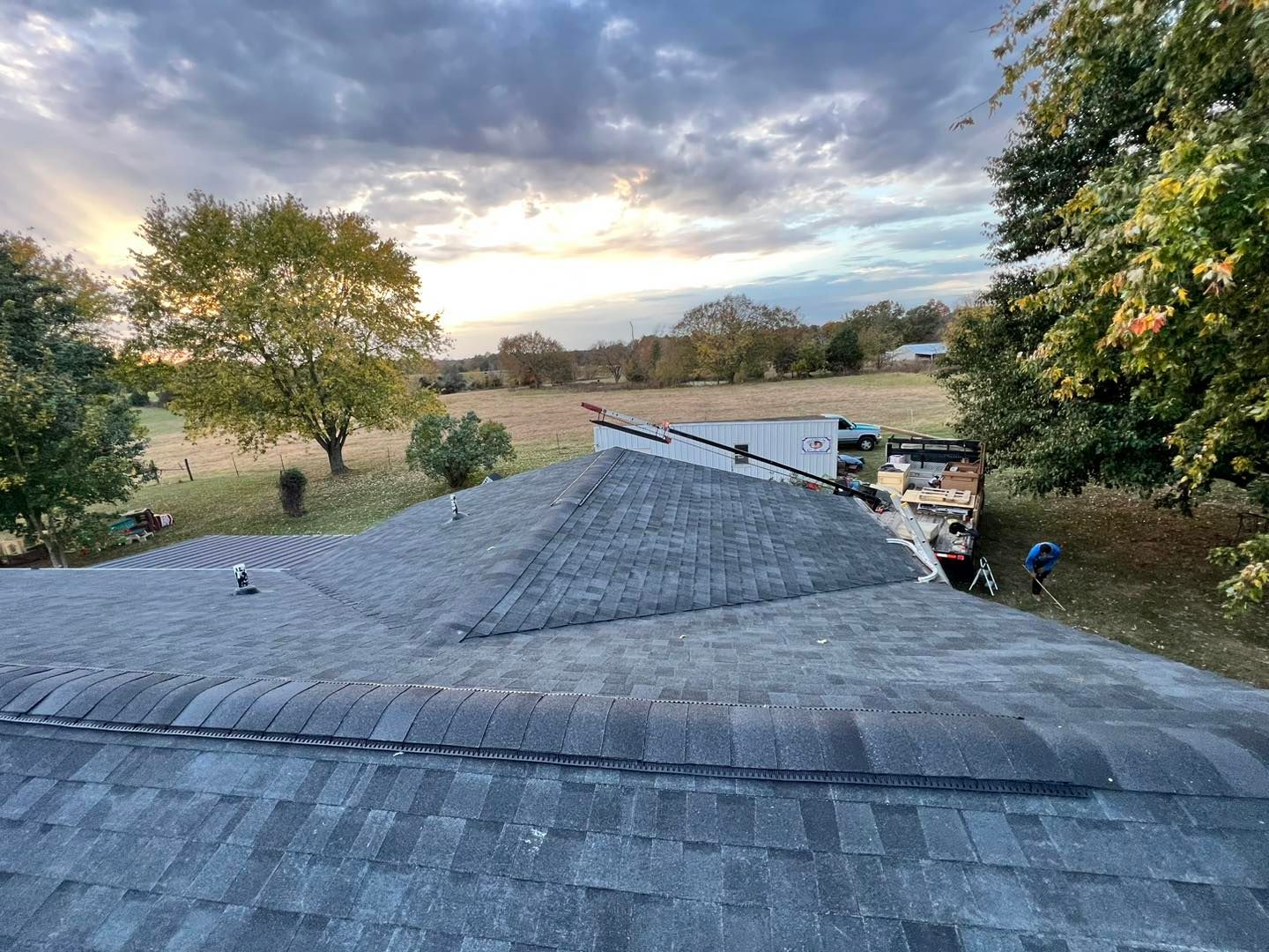 Overhead view of a roof with trees and a cloudy sky in the background; a white truck is in the distance.