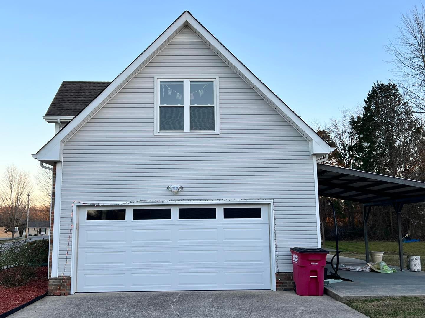 Two-story garage with white siding, white garage door, small window, and carport. A red trash can sits next to it.