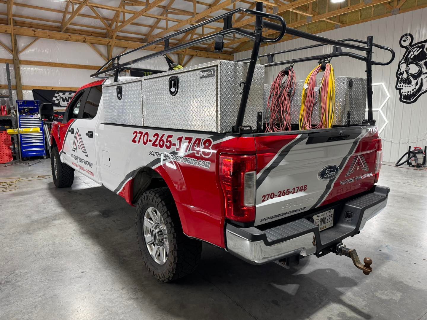 Red and white work truck with ladder rack, toolboxes, and company logo parked indoors.