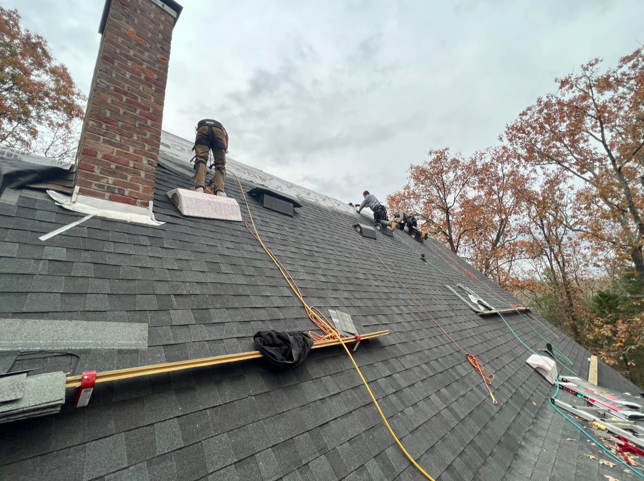 Roofers working on a dark gray shingled roof, near a brick chimney and fall trees.