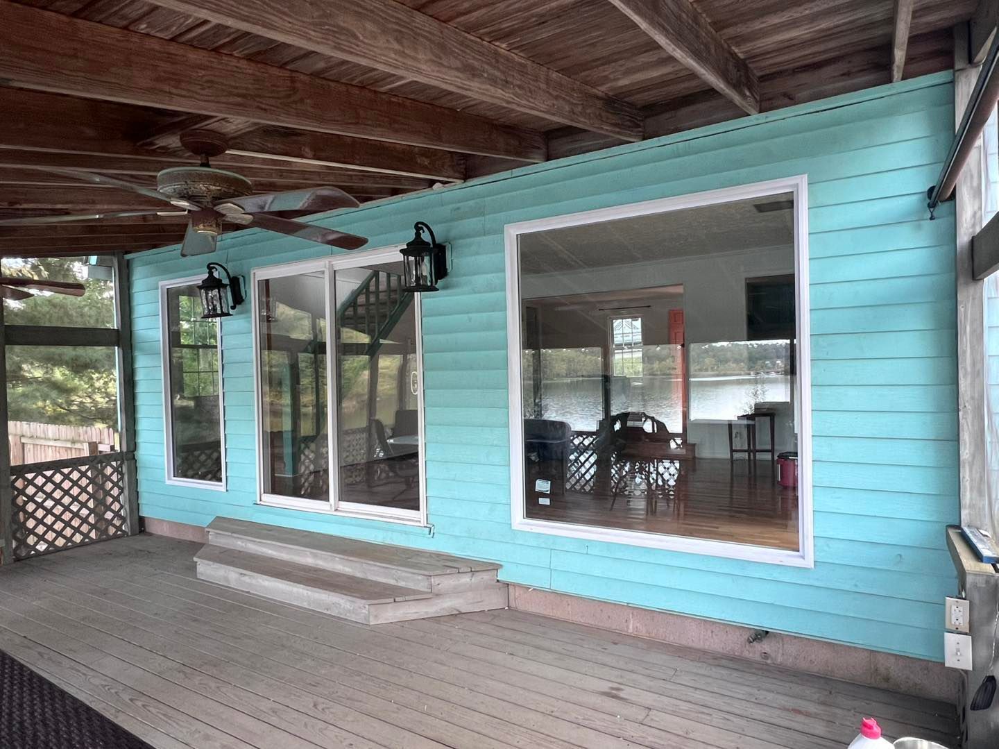 Covered porch with teal siding and large windows, reflecting interior. Wooden deck and ceiling.