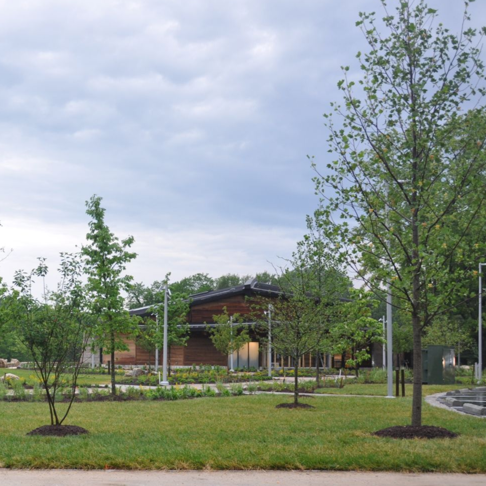 Building with glass walls, curving paved path with people biking, grassy field, and trees at sunset.