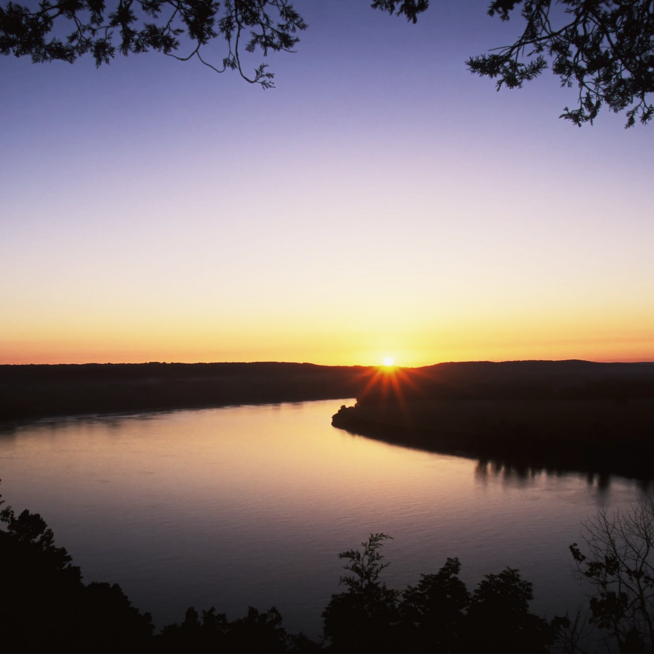 Sunset over a calm river, with silhouetted trees framing the horizon and sky.
