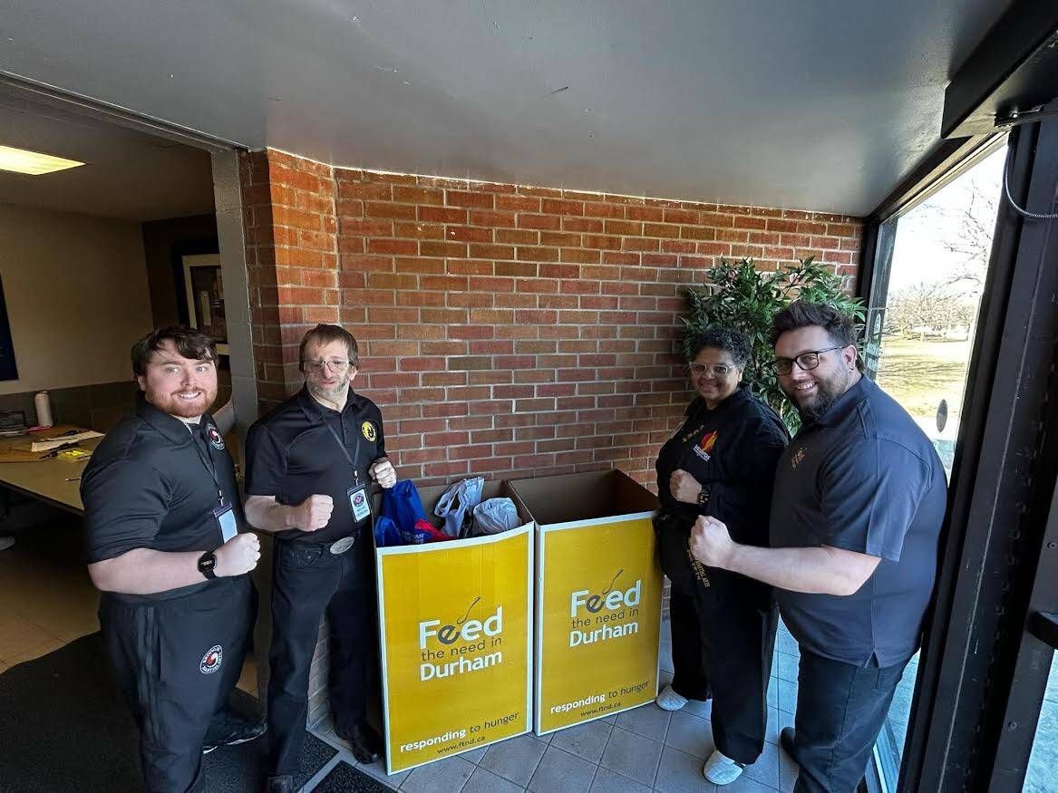 Four people stand on a porch beside yellow food donation bins against a brick wall.