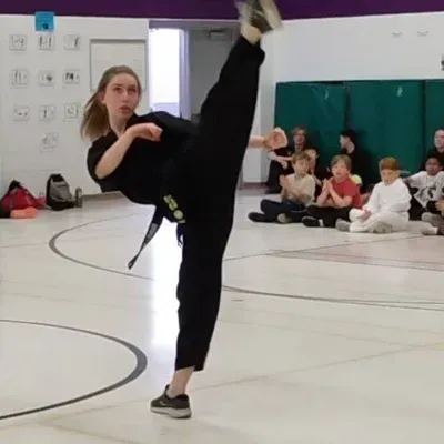 Martial arts student in black uniform performs a high kick in a gym while classmates watch.