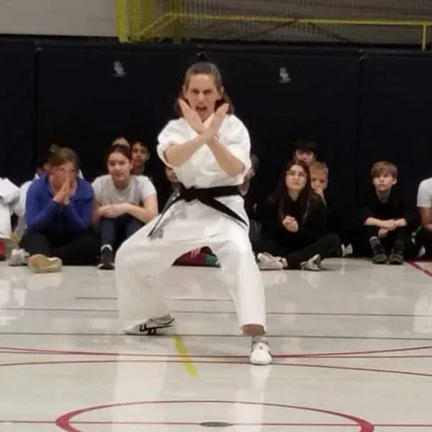Martial artist in white uniform performs a stance with crossed arms on an indoor gym floor.