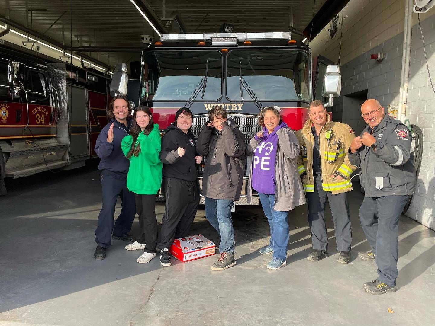 Group of people posing in front of a fire truck inside a garage, some holding gear and smiling.