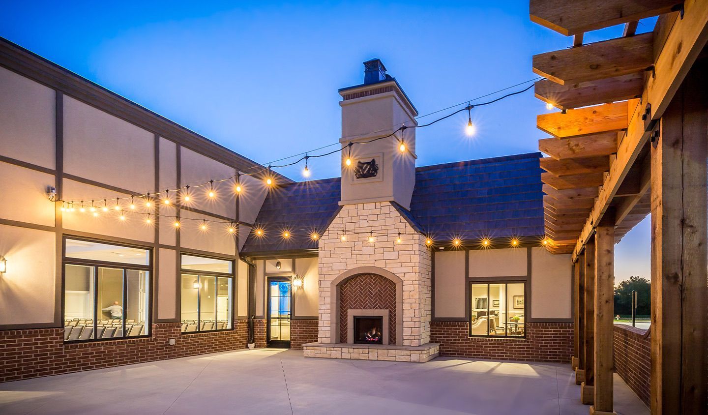 Modern courtyard patio at dusk with string lights, stucco walls, and a stone fireplace under a blue sky
