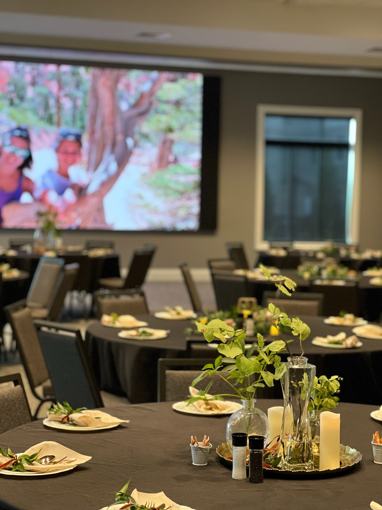 Banquet hall with set tables and plates, a large projected screen, and a floral centerpiece in the foreground