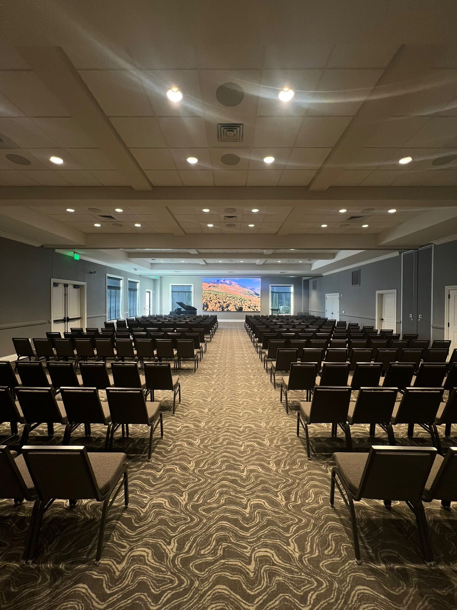 Empty conference room with rows of black chairs facing a stage and screen under recessed ceiling lights.