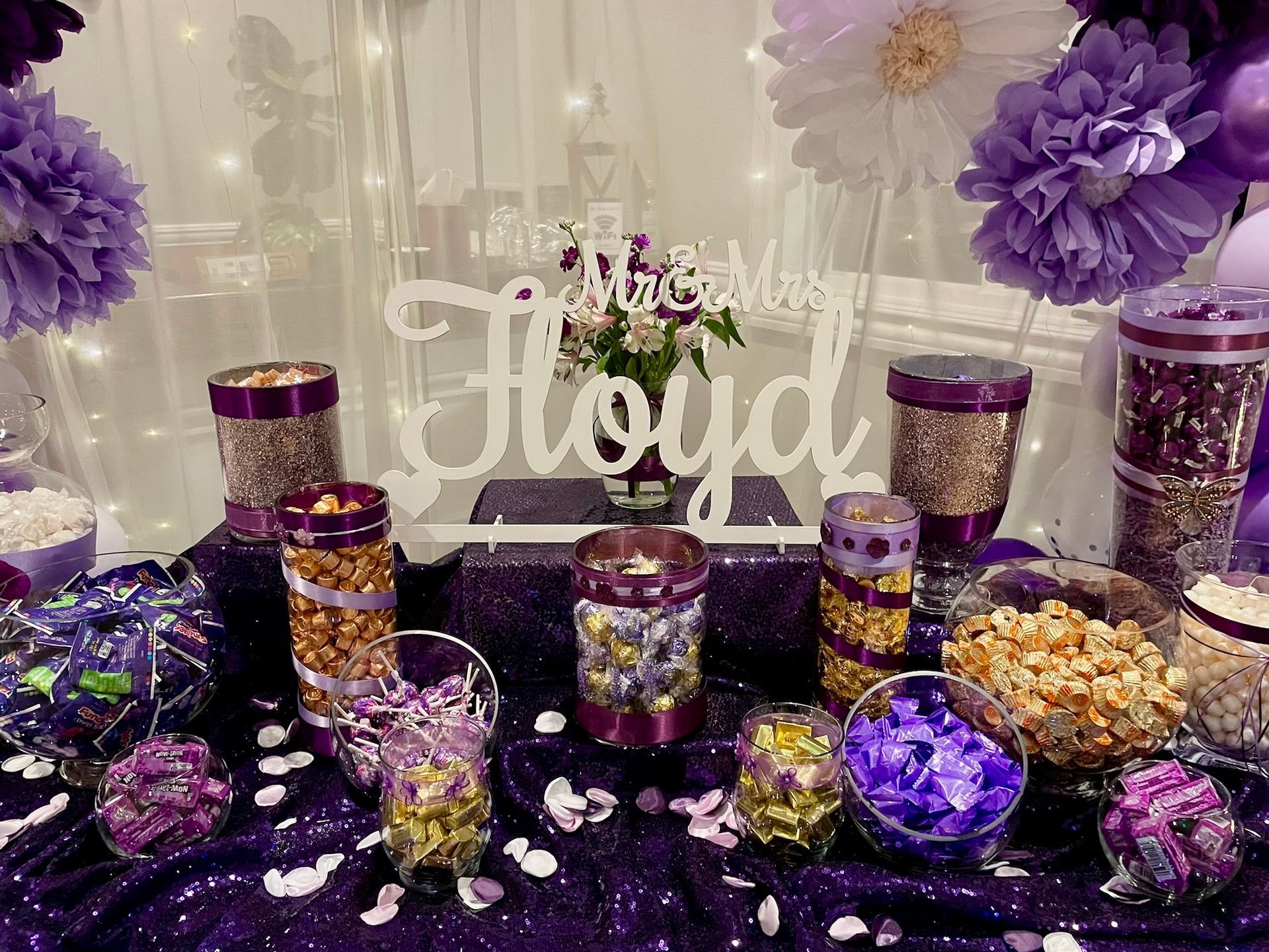 Purple and gold dessert table with jars, candy, and a “Mia Floyd” sign in front of floral decor