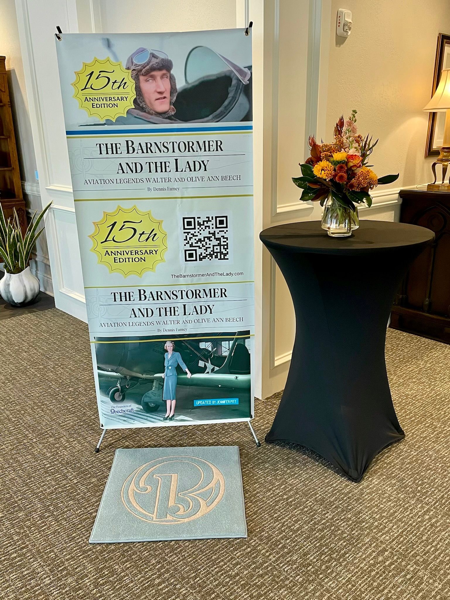 Event banner beside cocktail table with flowers, featuring a QR code and swimmer photos in a hotel lobby.