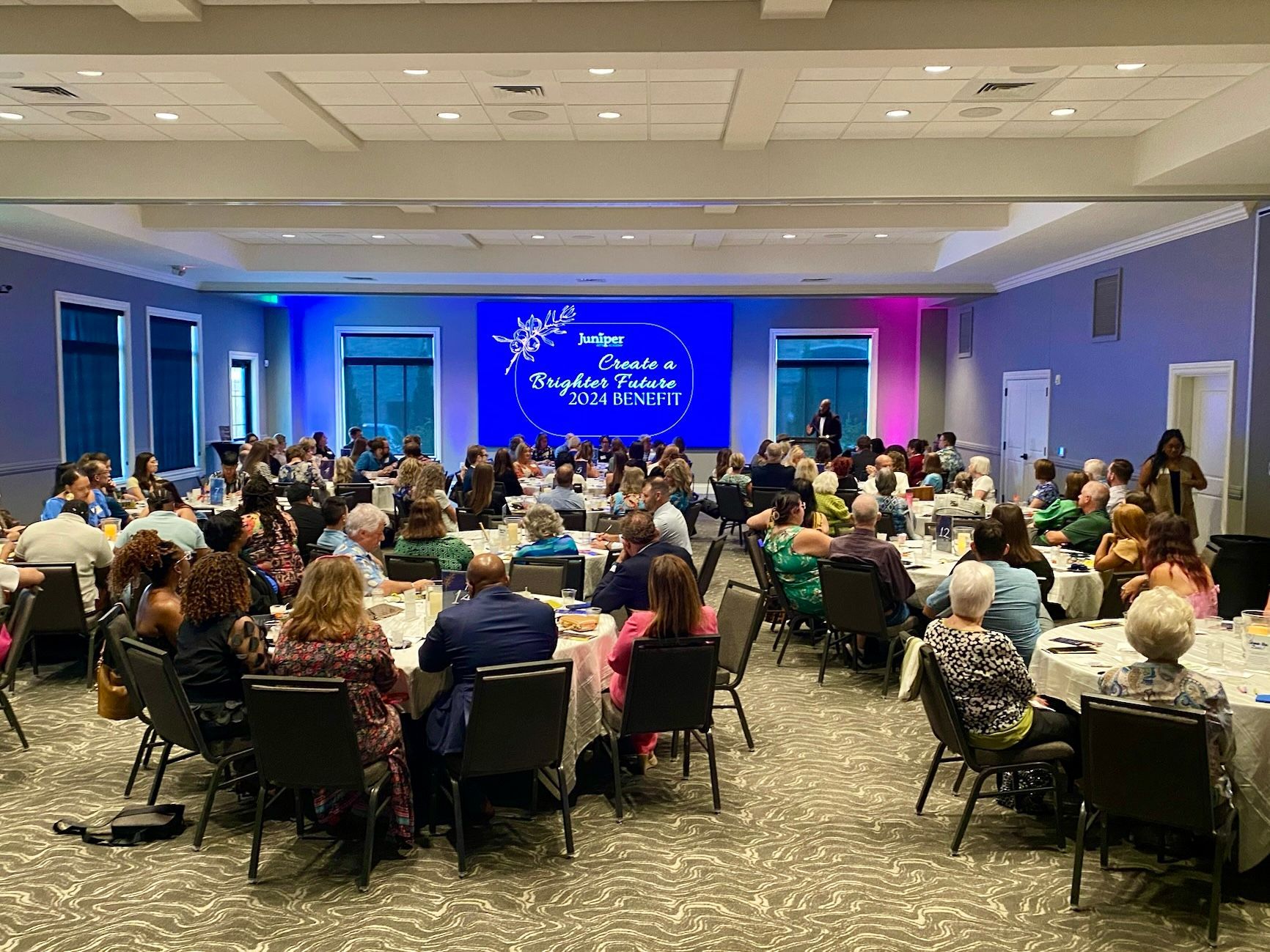 Conference audience seated in a large meeting room facing a blue-lit stage with a presentation screen