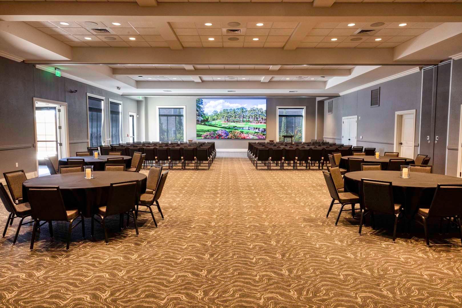 Spacious banquet hall with round tables and chairs, patterned carpet, and a large screen at the front.