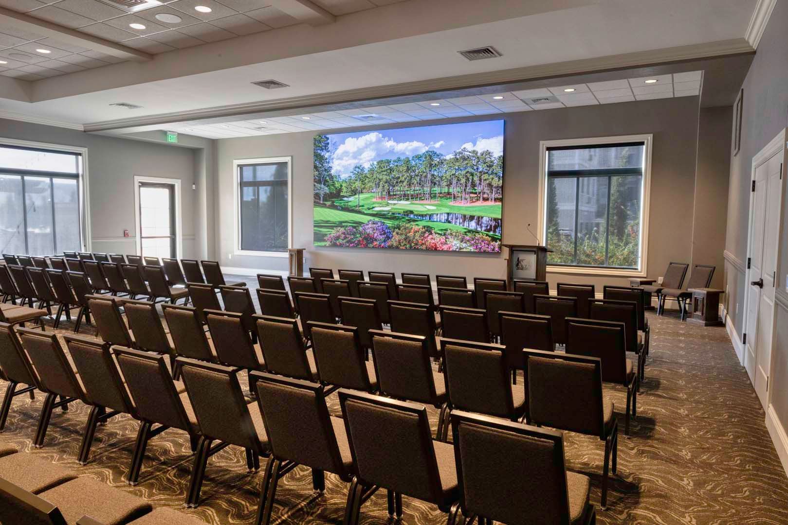 Empty conference room with rows of chairs facing a large wall-mounted screen showing a colorful landscape.