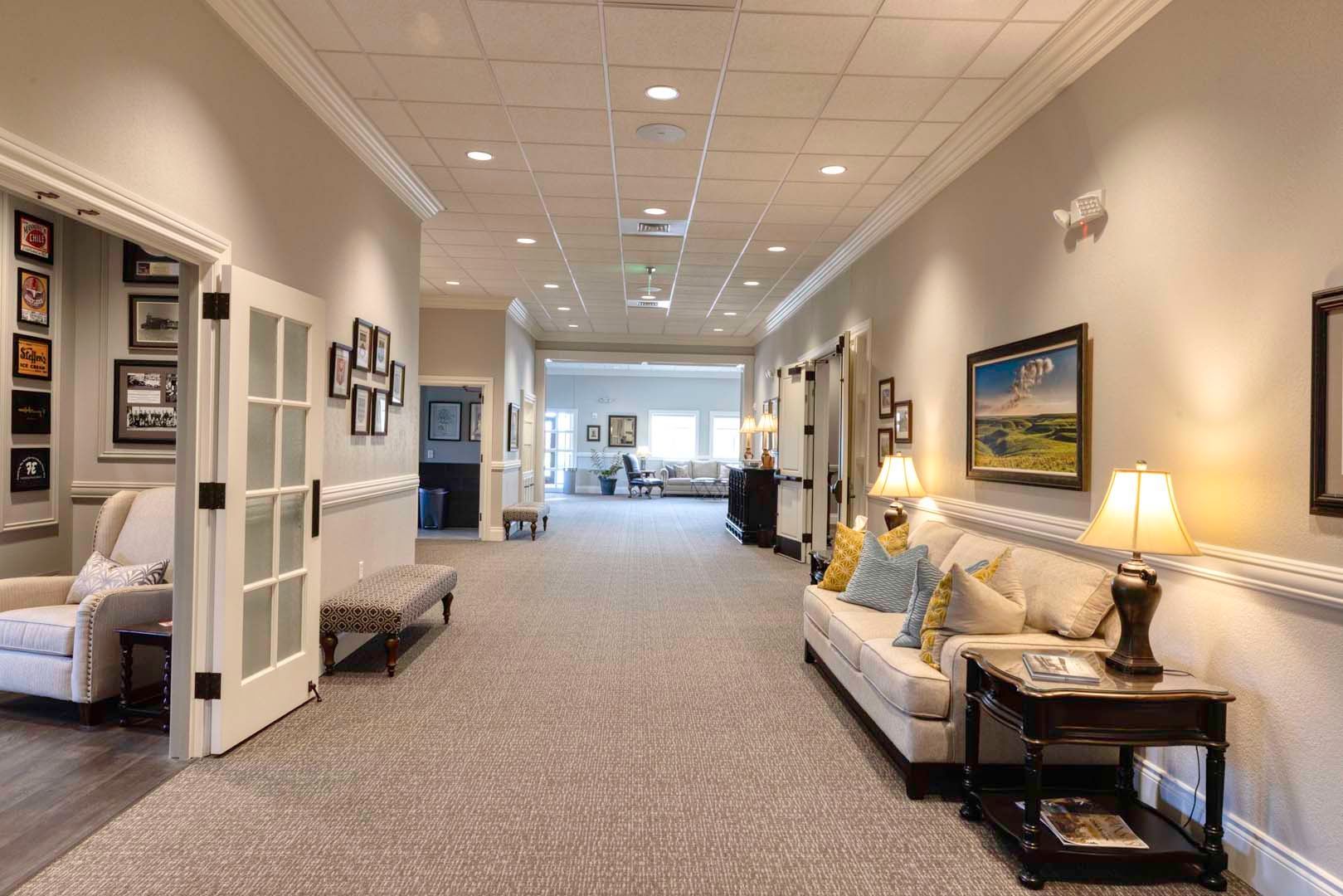 Bright hallway lounge with beige sofas, lamps, framed art, and carpeted floor leading to a bright windowed area