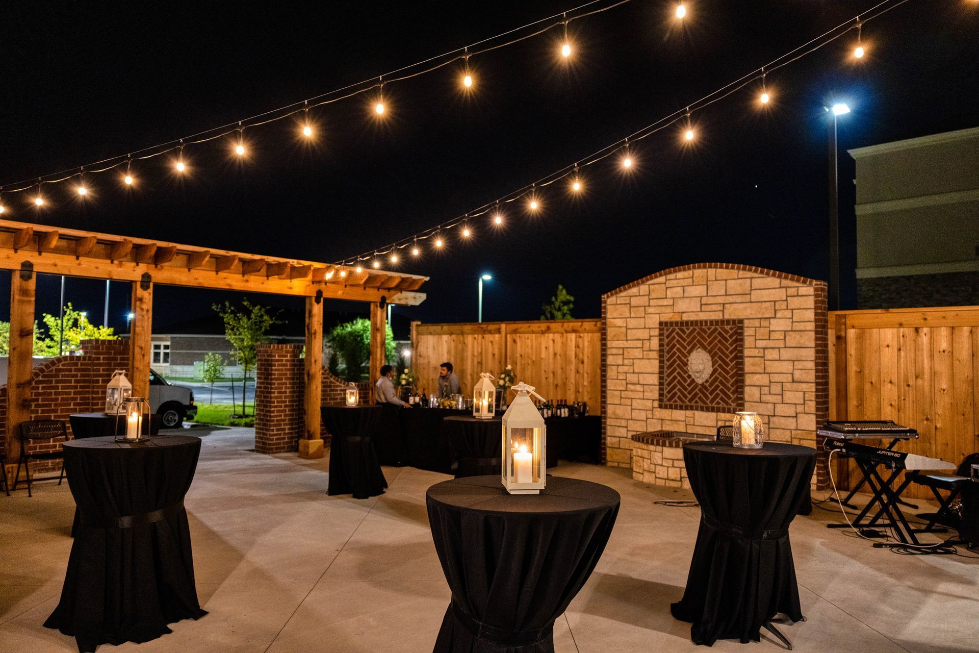 String-lit outdoor patio with high-top tables, lanterns, and a brick fireplace at night