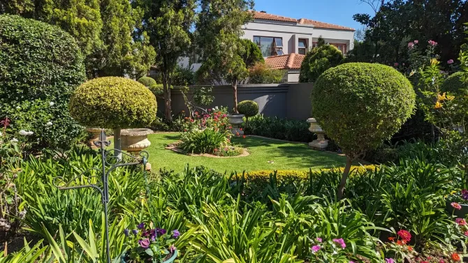 Lush green garden with trimmed bushes, flowers, and a house in the background under a bright sky.
