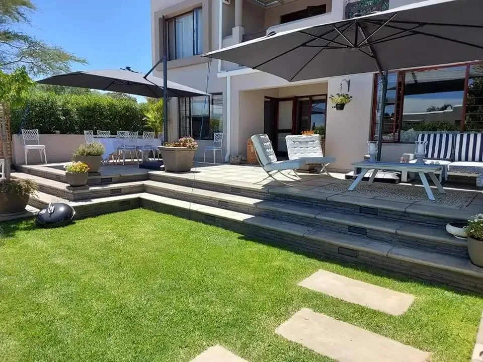 Backyard patio with steps, green lawn, umbrellas, seating, and a two-story house.
