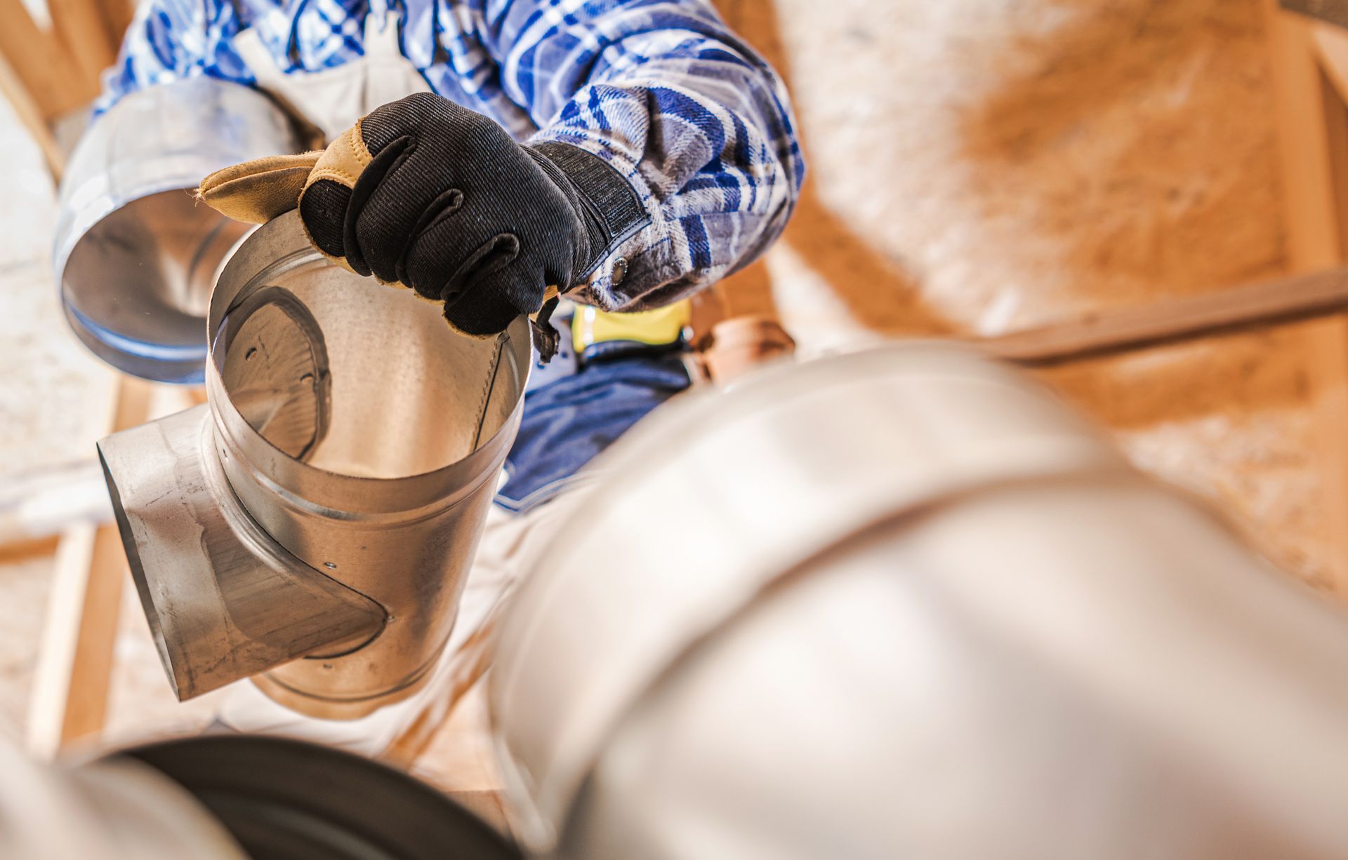 Person in gloves installing metal ductwork in an attic.