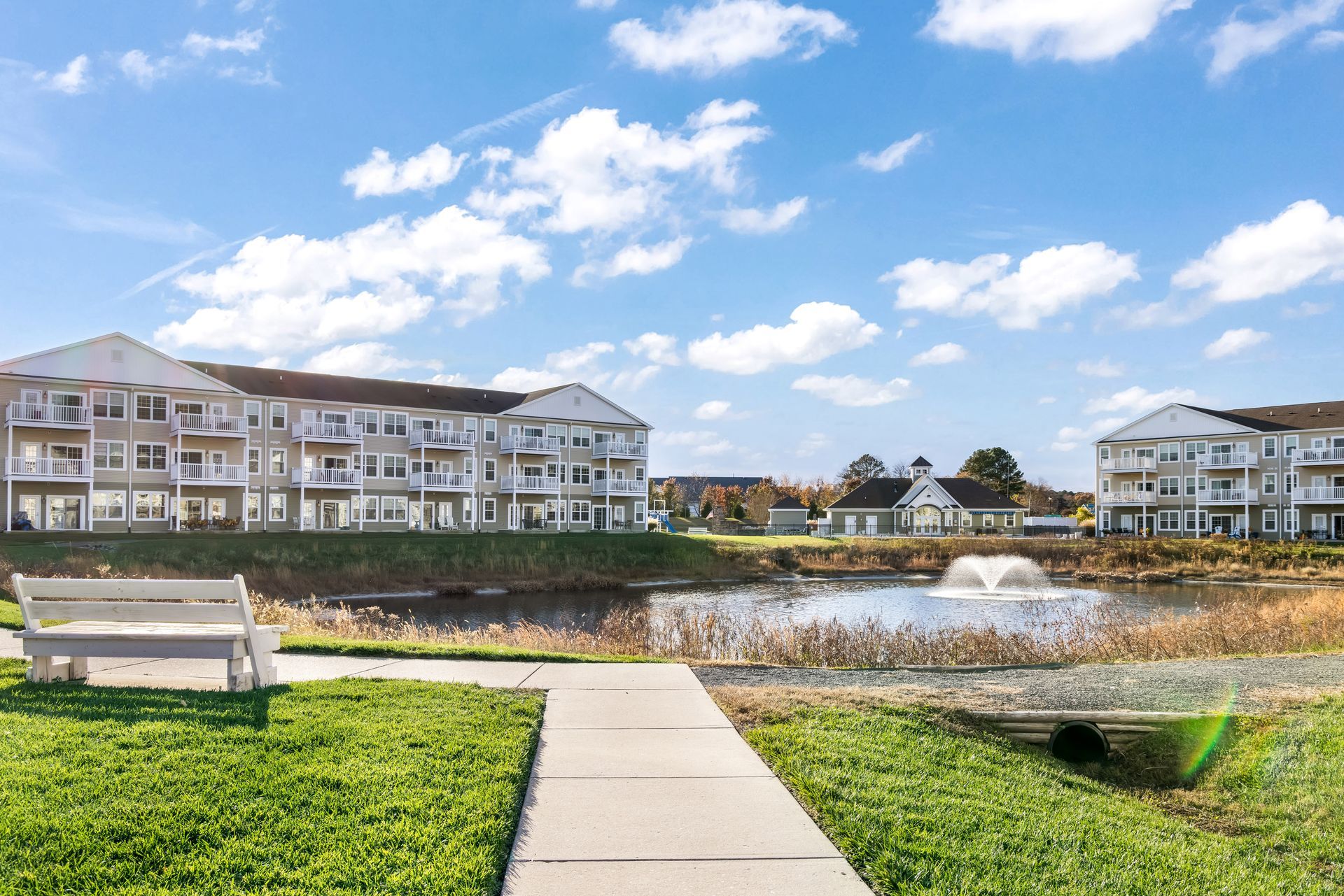 A white bench faces a pond and fountain between two multi-story apartment buildings under a blue, cloudy sky.