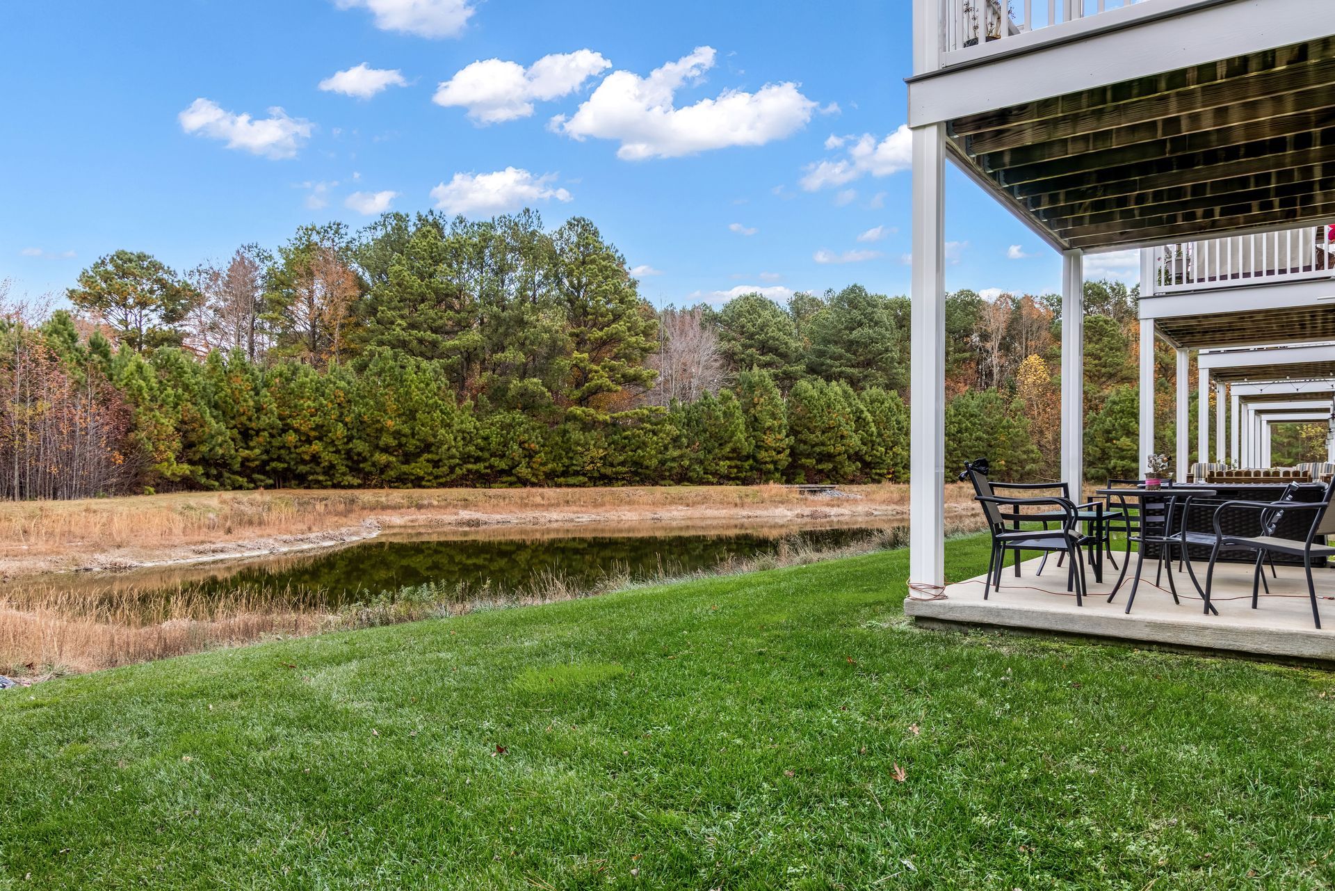 A view of a grassy yard with a small pond leading to a treeline next to a covered patio with outdoor furniture.