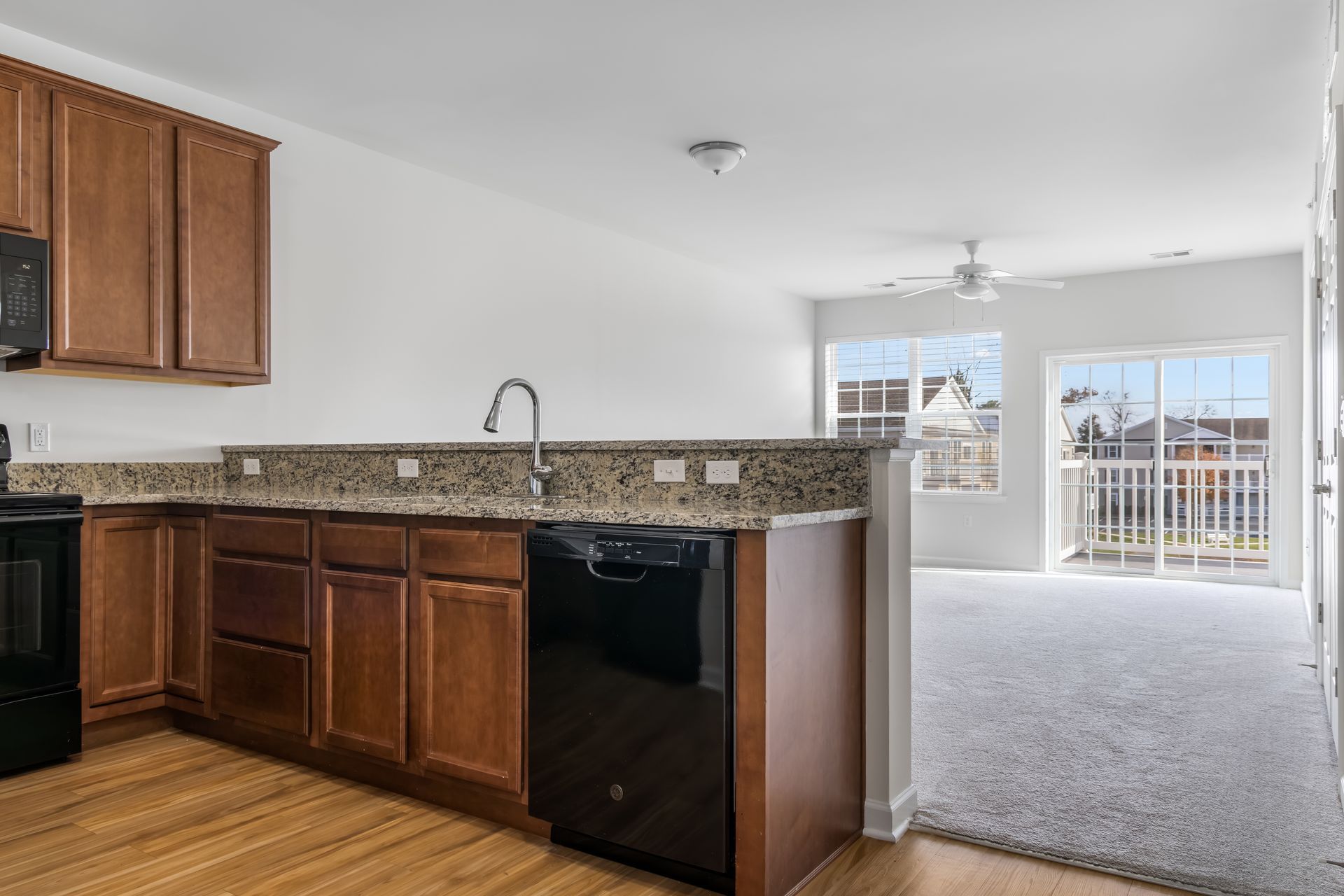A kitchen with brown wooden cabinets and granite countertops next to a carpeted living area with a sliding glass door.