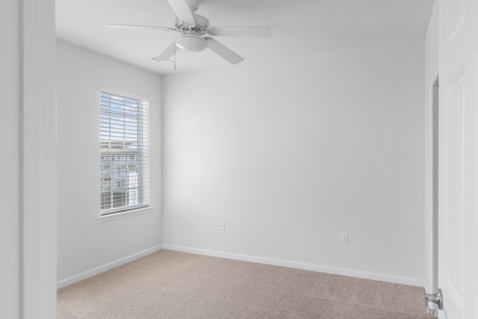 An empty bedroom with white walls, tan carpet, a ceiling fan, and a single window with white blinds.