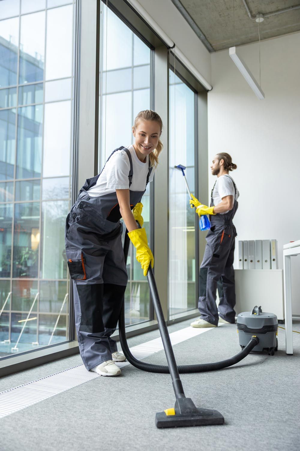 Two cleaners vacuuming a bright office with large windows and a carpeted floor Two cleaners vacuuming a bright office with large windows and a carpeted floor