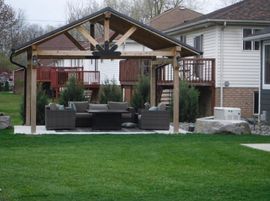 Covered backyard patio with outdoor seating and a wooden roof beside a house.