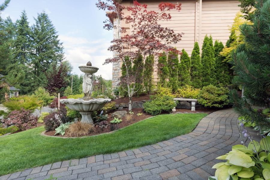Landscaped garden with stone fountain, curved paver path, shrubs, and a house exterior