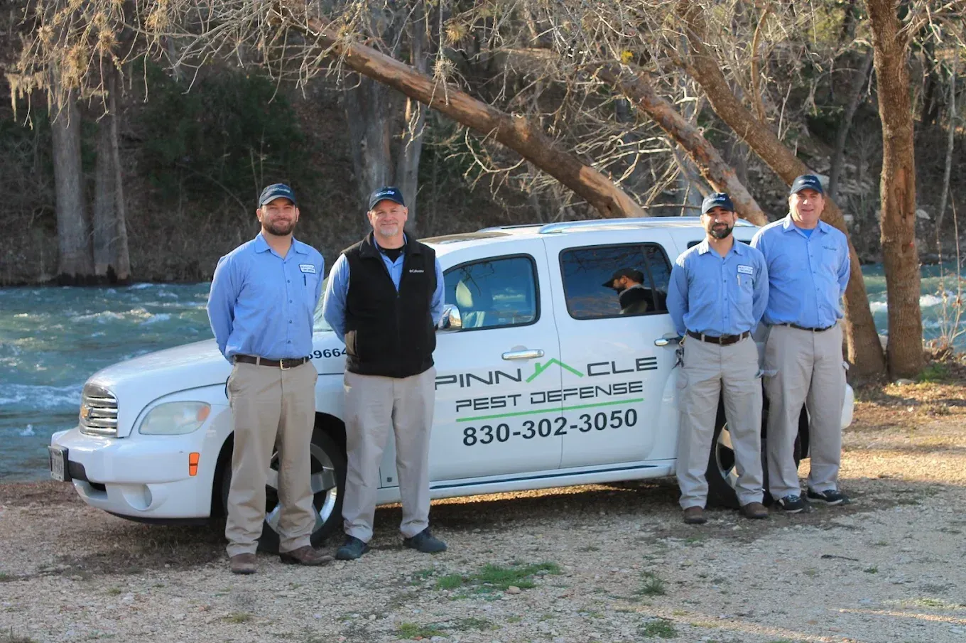 Six people in gray uniforms posing outdoors by a lake with trees and a large tree trunk.