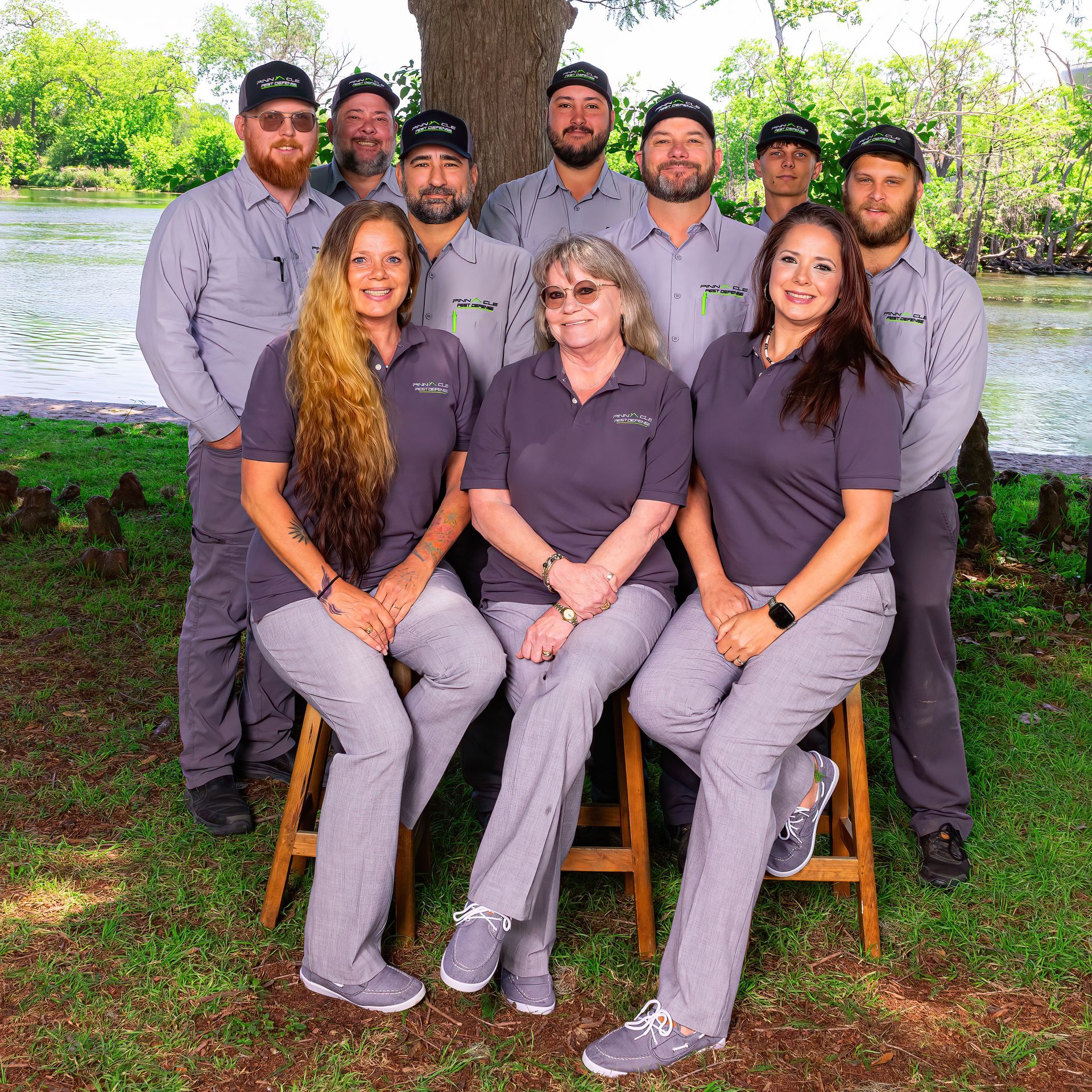 Seven people in gray uniforms posing outdoors by a lake and tree, with three seated in front and four standing behind.
