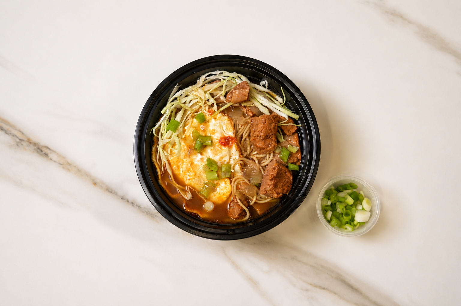Black bowl of noodle soup with vegetables, topped with herbs, beside a small cup of chopped green onions.