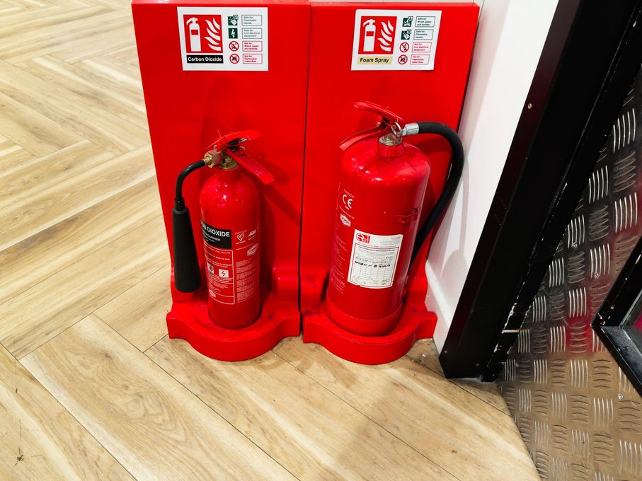 Two red fire extinguishers on a wooden floor beside a wall and black panel.
